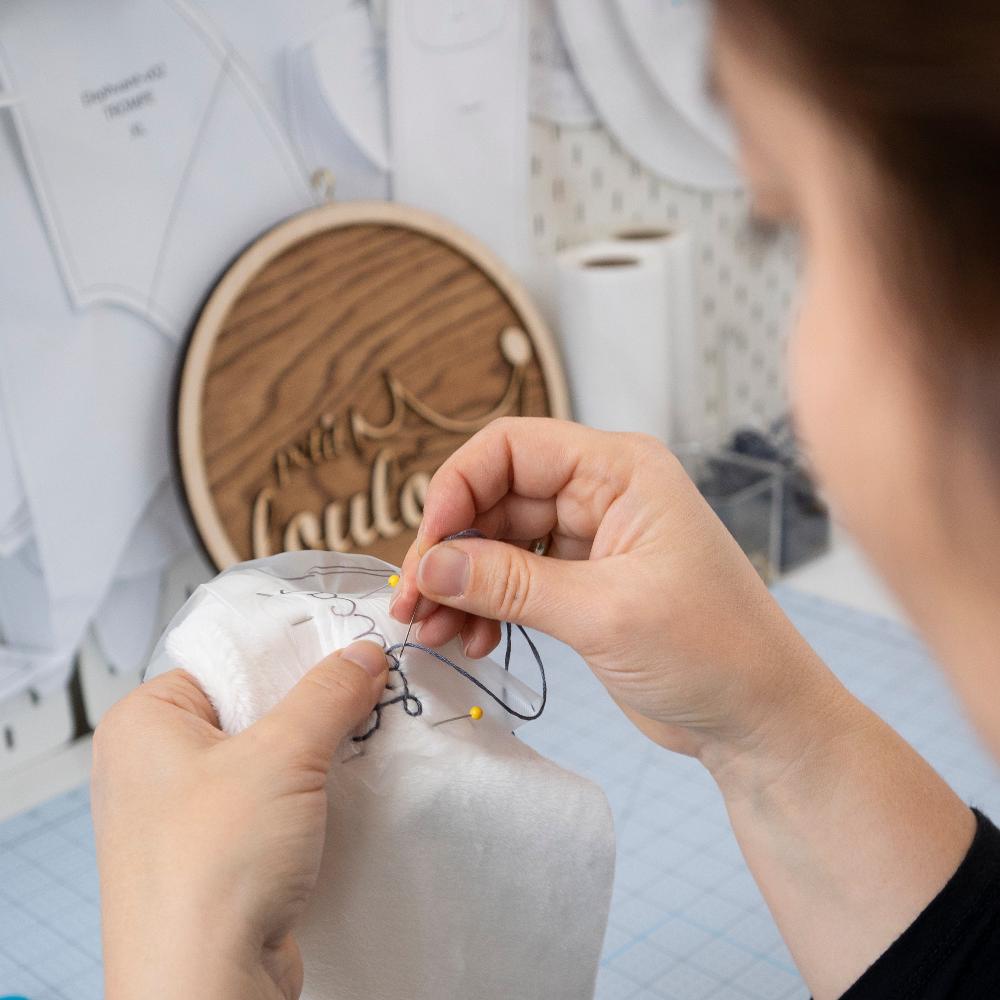 Close up of hands sewing a plush toy, in the background you can see a wooden signs that reads "petit Loulou"