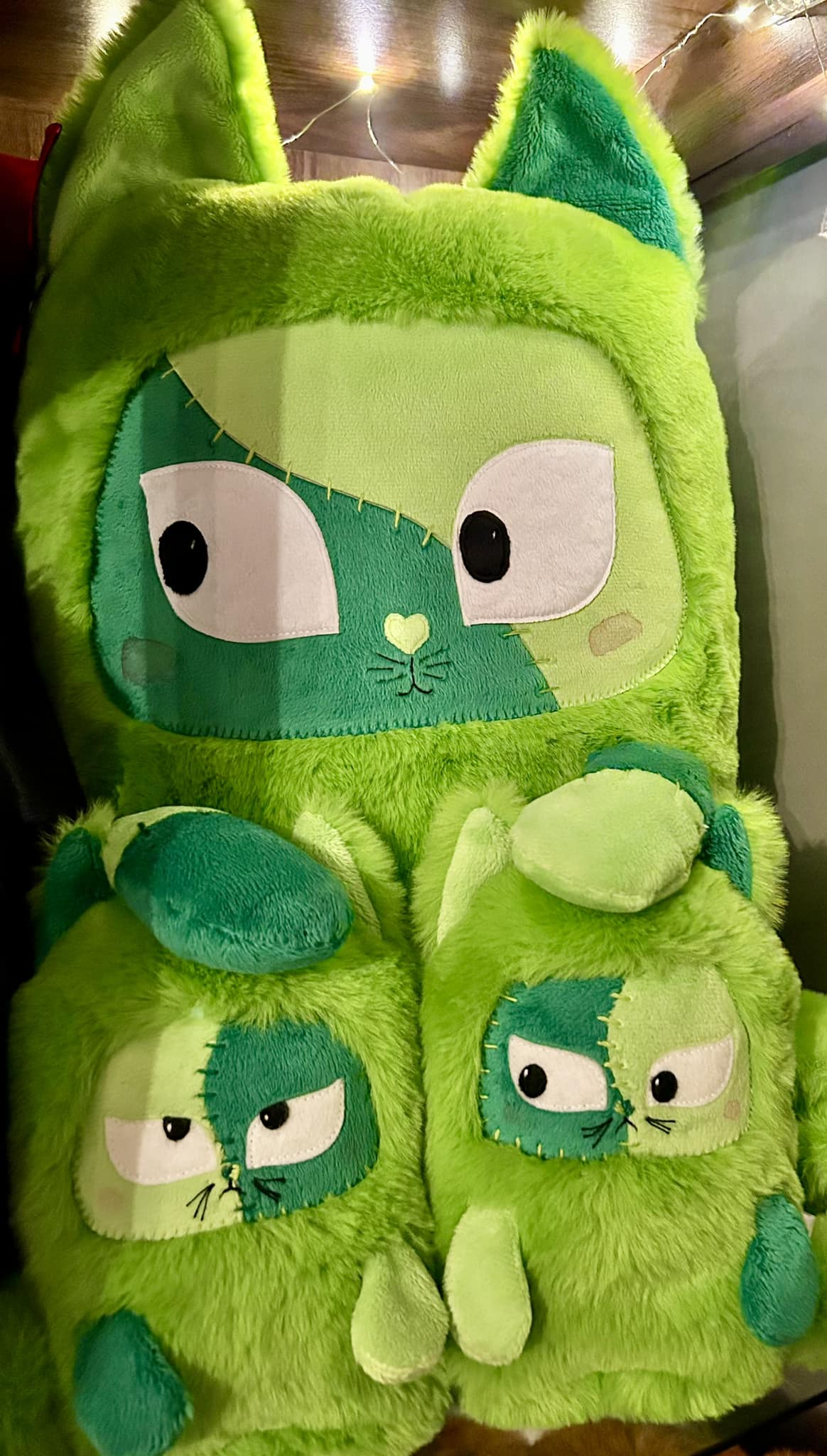 A large green cat plushies with his stitched face has his paws on 2 smaller green grumpy cat plushies. They all look like Frankenstein